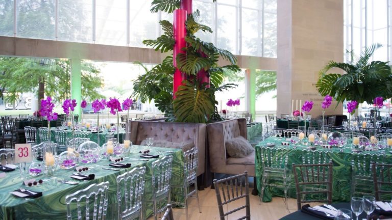 Daytime event setup at the Meyerson inspired by island life, purple orchids, palm leaves, pink and glass accents on rectangle tables