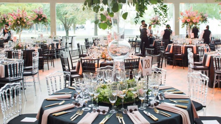 Sofisticadas y elegantes mesas negras y rosas con mesas redondas, centros de mesa de gran tamaño en el vestíbulo de Meyerson - un grupo de servidores se reúnen en la parte trasera haciendo preparativos de última hora