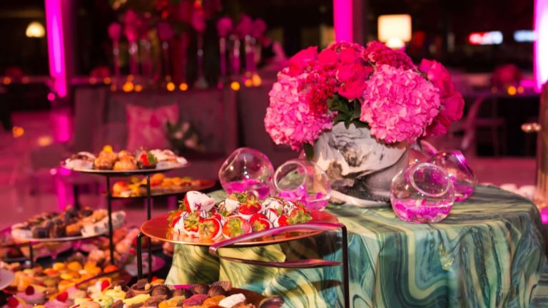Dramática barra de postres con trufas, fresas cubiertas de chocolate, macarrones, tartaletas y más con dramáticas luces rosas y arreglos florales.