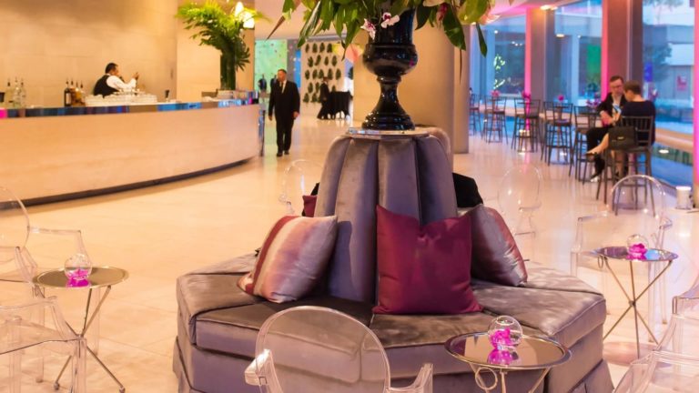 Zona de asientos con banquetes circulares, flores y sillas fantasma en una romántica paleta rosada.