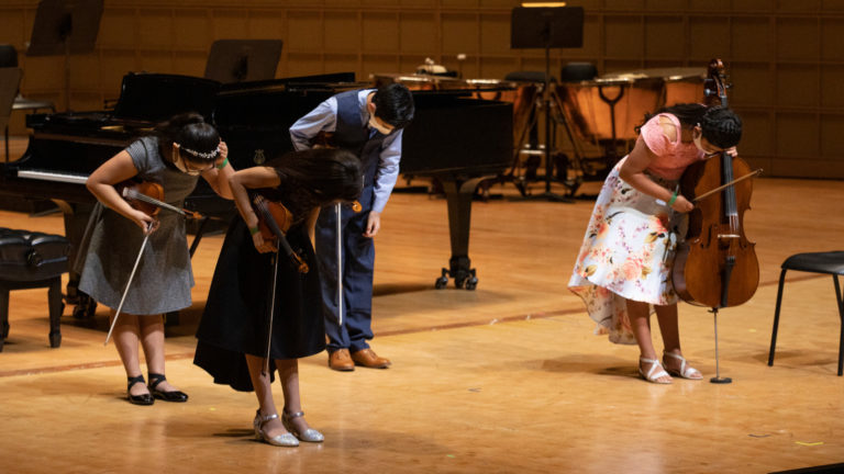 Four children bow on stage holding instruments