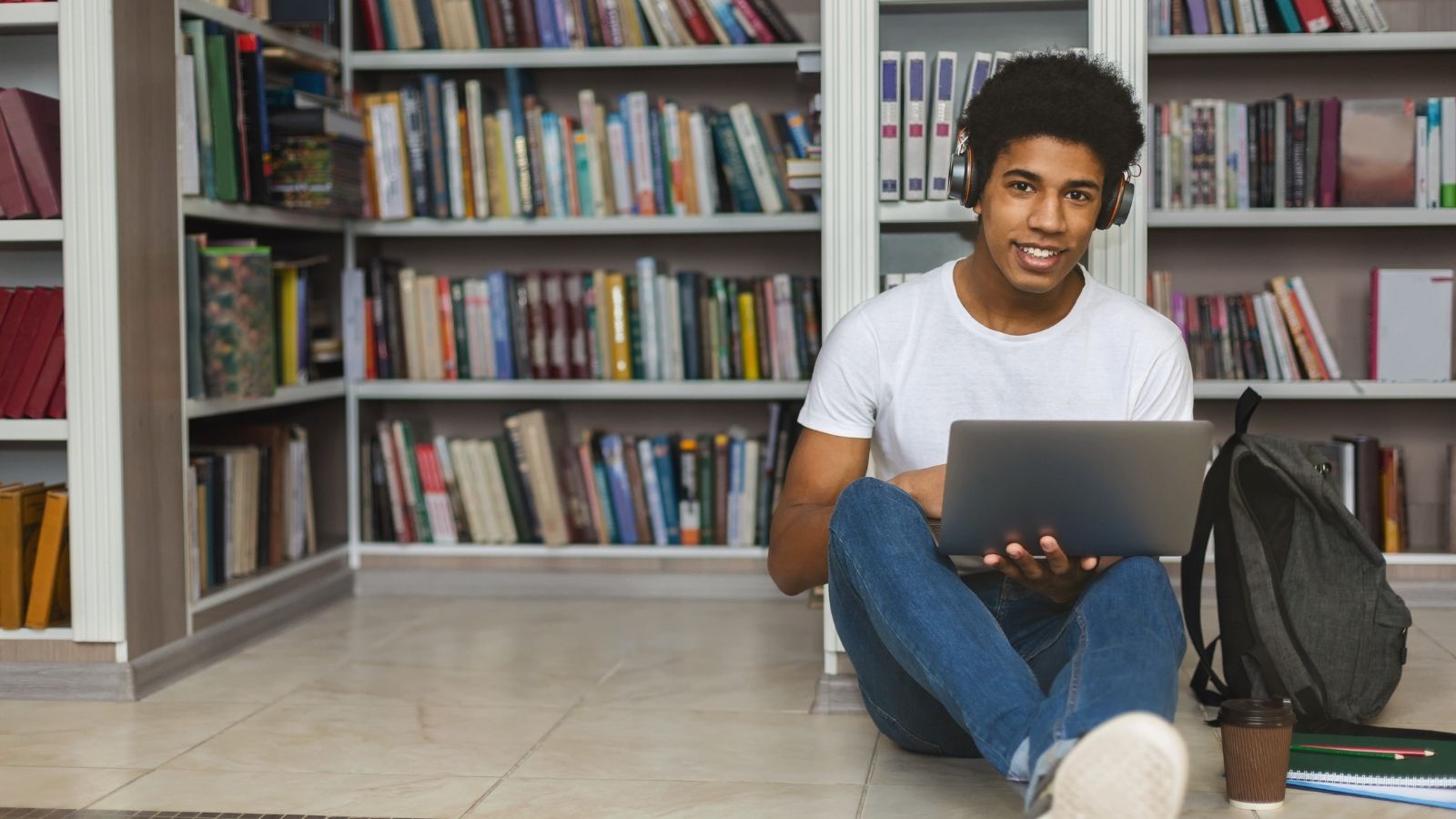 Hombre universitario sentado en el suelo de la biblioteca escuchando los auriculares y trabajando en un portátil